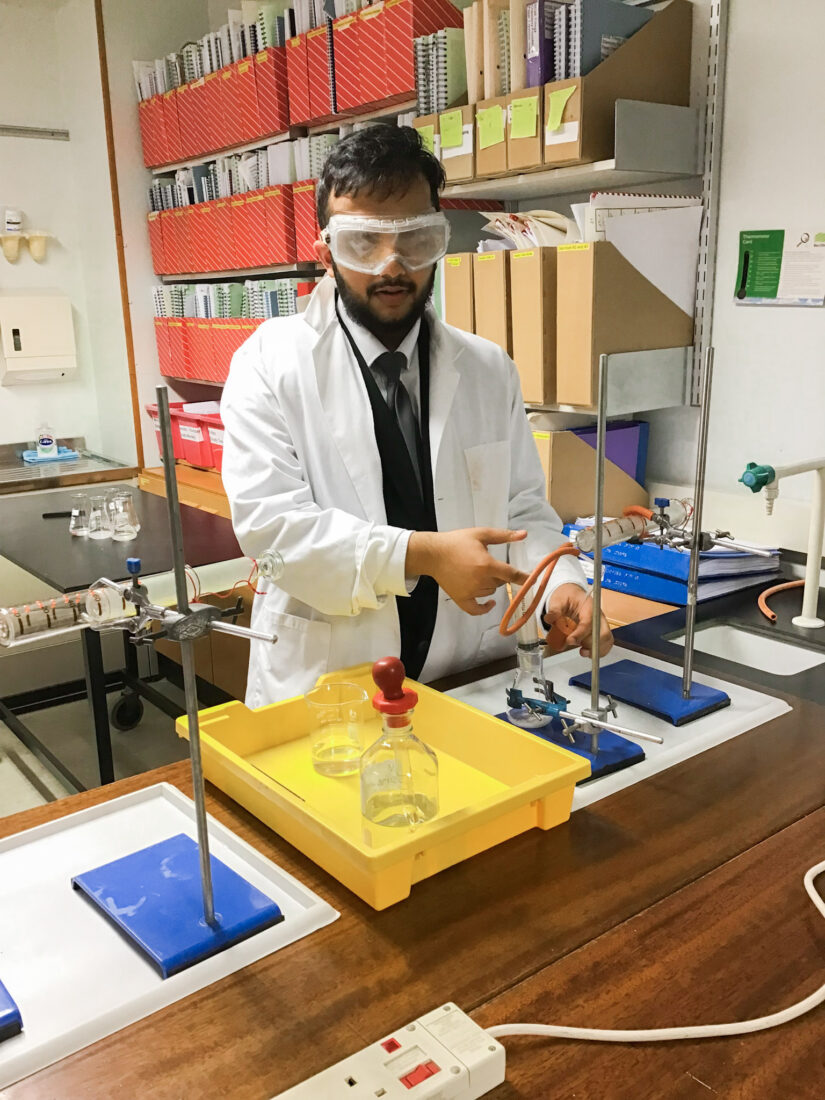 Male VI student in white laboratory coat in the Science room. He is using an adapted syringe to measure out acid and adding it to calcium carbonate
