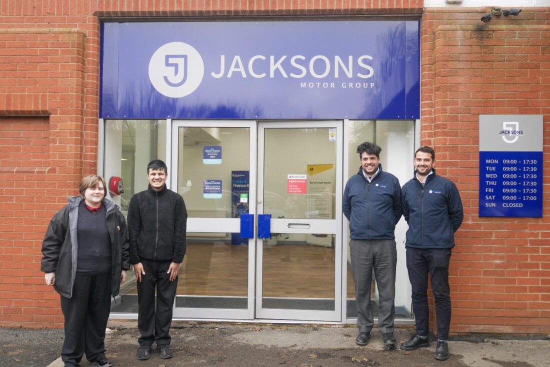 Picture of two students and Adam and Sher Jackson outside the garage. There is a large blue logo for Jacksons above the door. Everyone is looking at the camera smiling.
