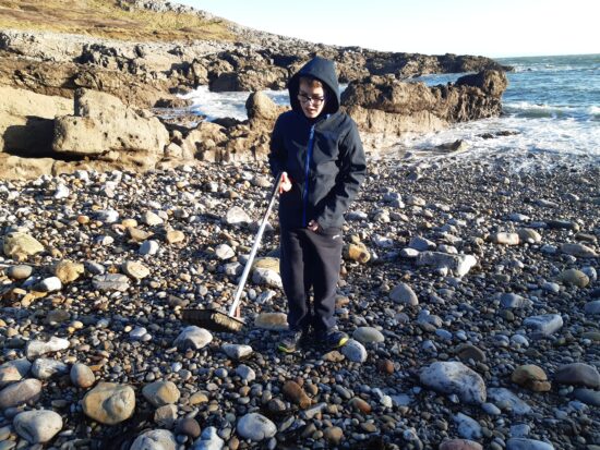image is of pupil on the beach with a litter picker. The beach is pebbled and the sea is in the background.