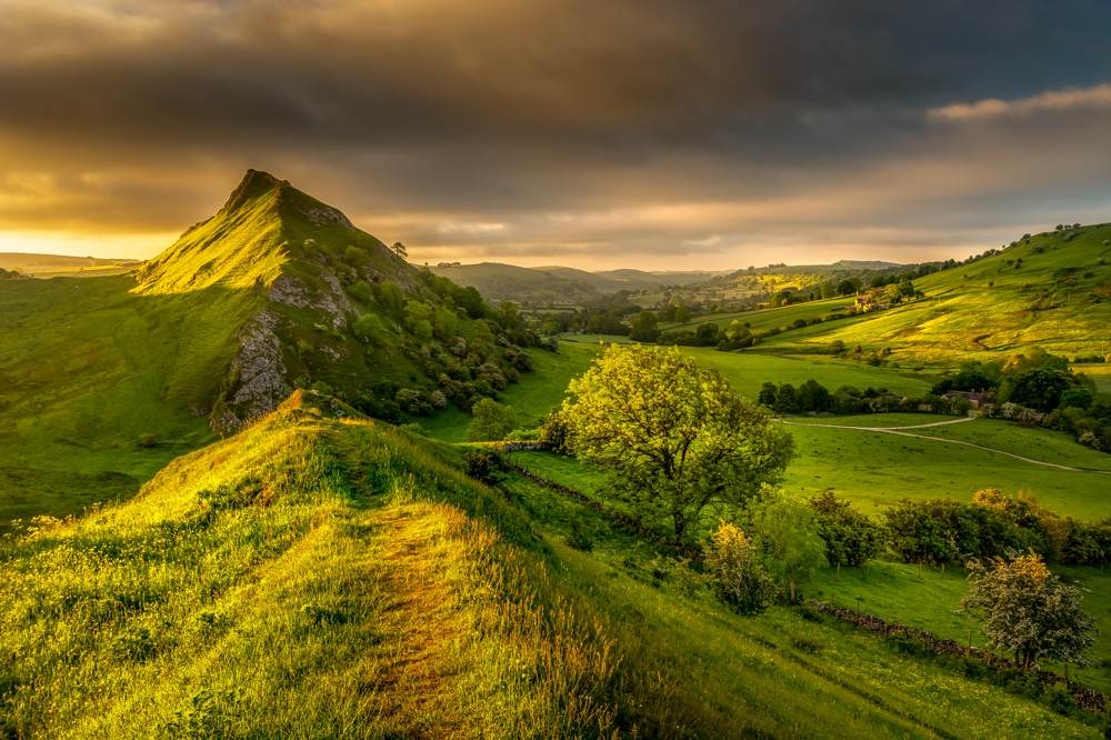 image is of the Peak District Hills. The grass is very green and lush. The sky is dramatic with lots of clouds. There are rays of sunshine coming through the clouds.