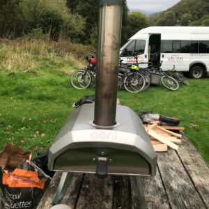 Pizza stove in use on a picnic bench with bikes and the NCW minibus in the background