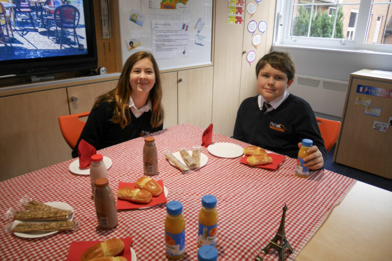 Students Amelie and Roman smiling at the camera, sat at the table with brioche and juice for their chosen breakfast