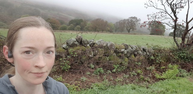 image of Naomi smiling at the camera. There is a stone wall and fields in the background.