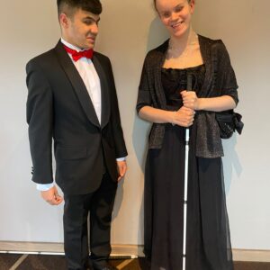 Mustafa is dressed in a black suit with red bow tie. Martha is in a black dress holding her cane. Both students are smiling at the camera