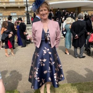 Rosie is wearing a blue dress and fascinator with a pink bolero. She is smiling at the camera