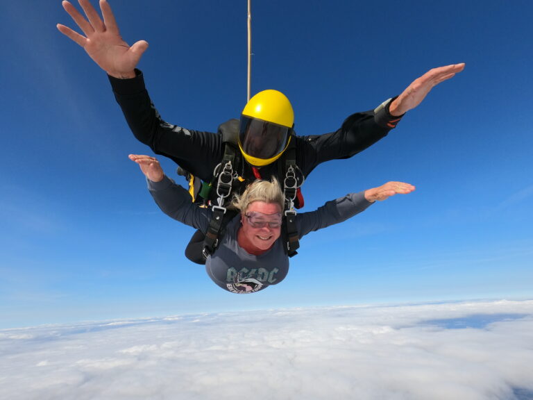 image of vicci in the air sky diving. She is attached to an instructor. The sky is very blue and they are above the clouds