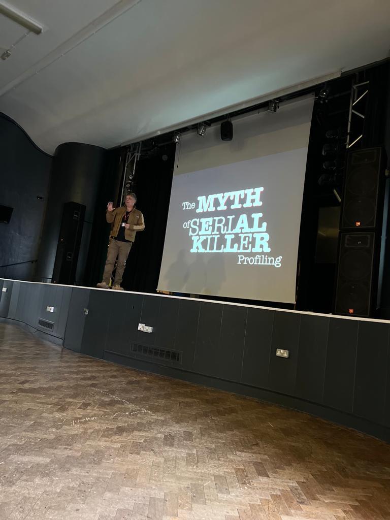image of a stage with a speaker and a slide showing the words 'Myths of a serial killer'