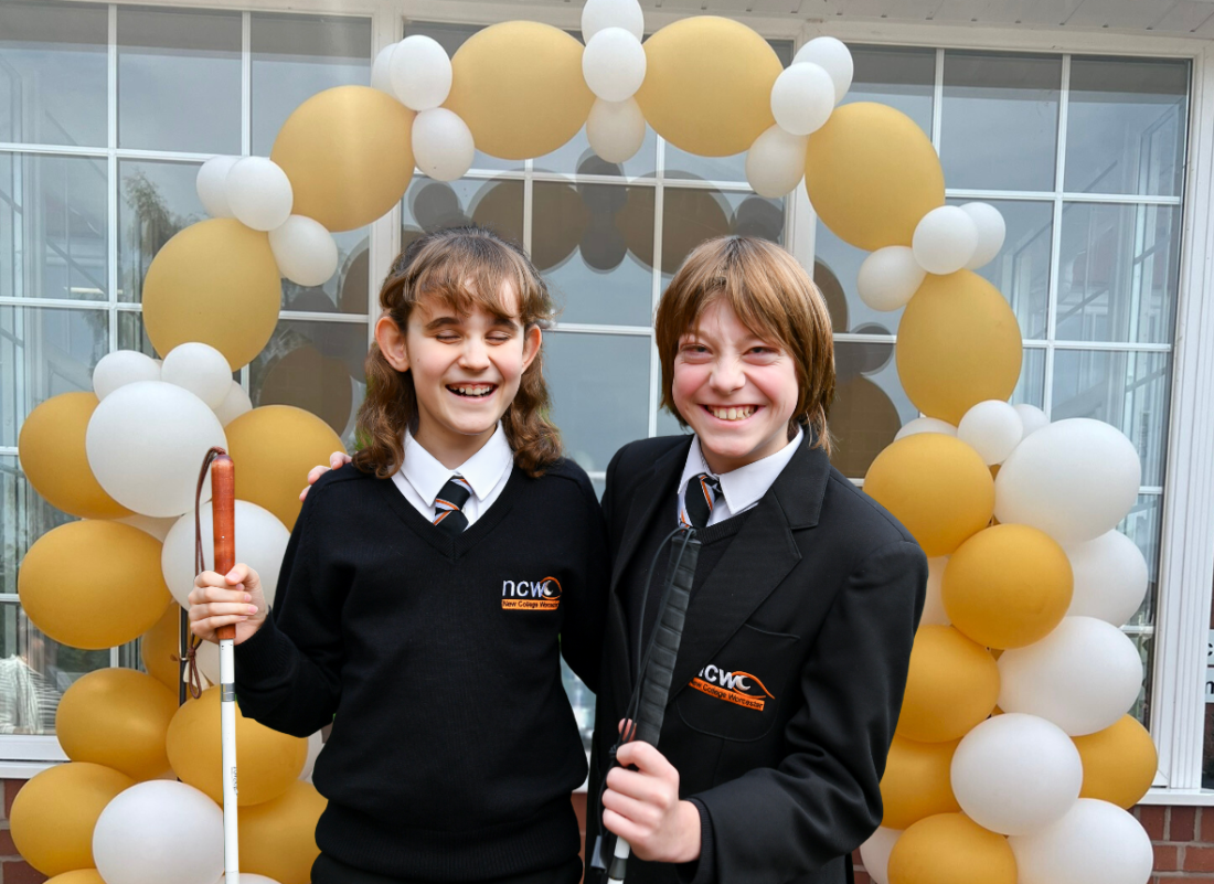 Male and female students smiling towards the camera with gold and pearl balloons in the background. Both are holding canes