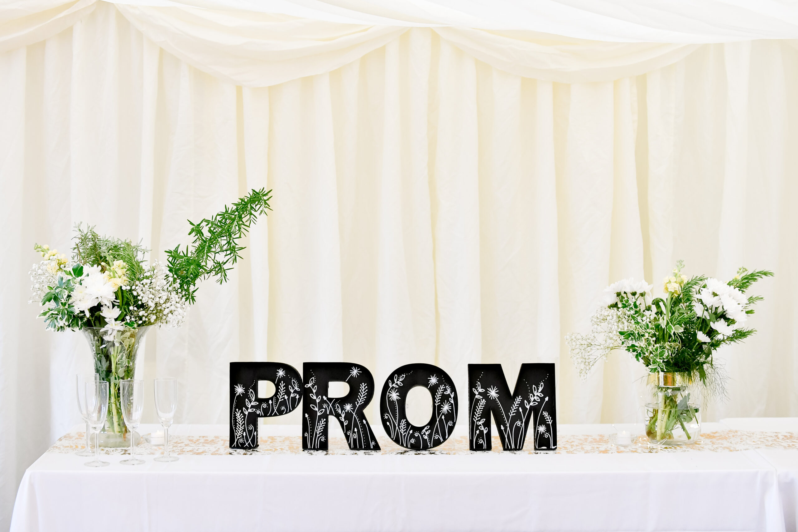 Photo of table with white table cloth with stand up letters saying prom. A vase of white and green flowers are either side of the letters
