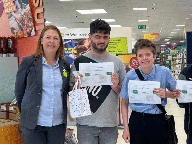 Two students smile as they receive a certificate from a member of the Waitrose team, celebrating the completion of their work experience.