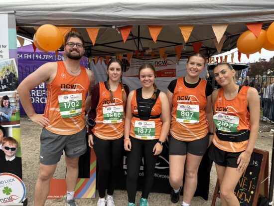 A group of 5 runners smiling for a group photo. They are wearing orange NCW running vests.