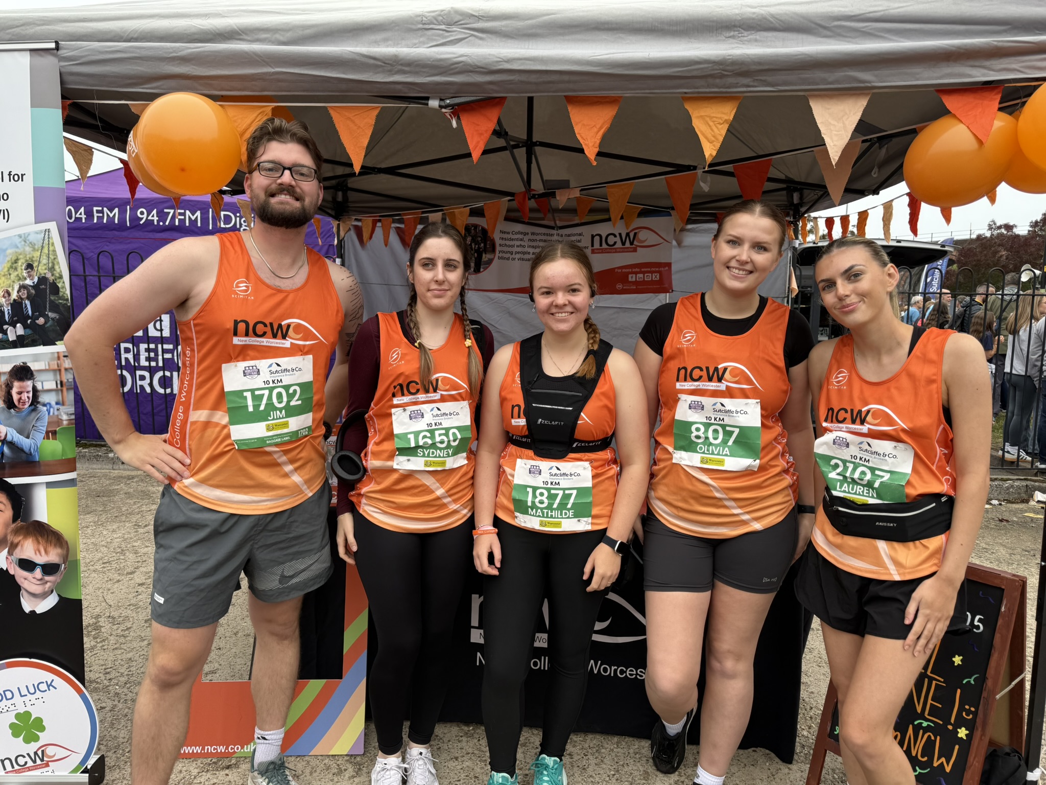 A group of 5 runners smiling for a group photo. They are wearing orange NCW running vests.