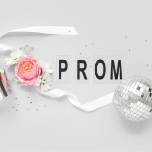 A flat‑lay arrangement featuring a silver paper crown, a pink rose with white flowers, a white ribbon, and a silver disco ball. The word “PROM” is spelled out in black letters on individual white cards against a light grey background, with scattered small silver confetti.