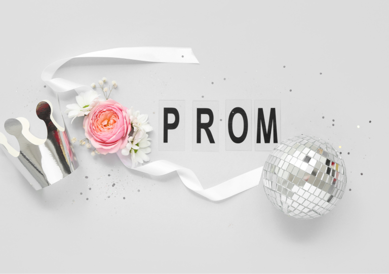 A flat‑lay arrangement featuring a silver paper crown, a pink rose with white flowers, a white ribbon, and a silver disco ball. The word “PROM” is spelled out in black letters on individual white cards against a light grey background, with scattered small silver confetti.