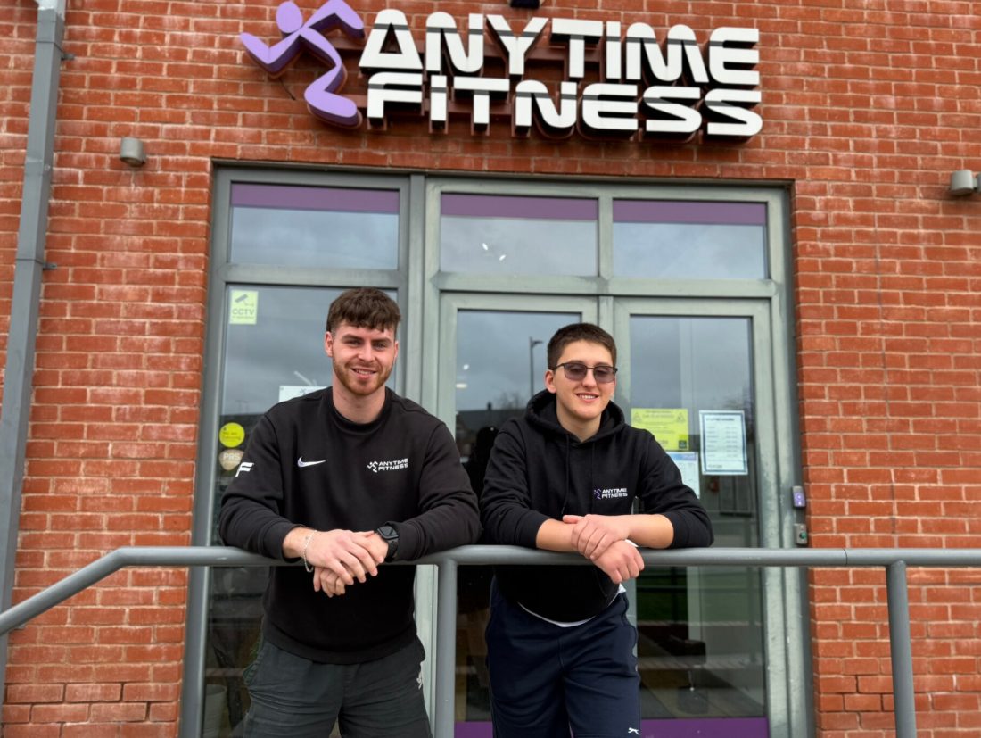 Dom and Ally standing outside the entrance of an Anytime Fitness gym, leaning against a metal railing in front of the building. Both individuals are wearing dark athletic clothing with the Anytime Fitness logo on the front. Behind them is a brick wall, large glass double doors, and a prominent Anytime Fitness sign in white and purple above the entrance.