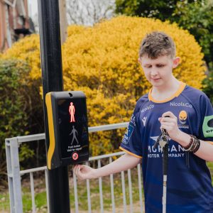 A student standing at a pedestrian crossing presses the button on the signal box with one hand while holding a white cane in the other. The signal box displays a red standing figure indicating not to cross. Behind the person is a metal railing, green shrubbery, and a large yellow bush. A building with red brick and white window frames is visible to the left.
