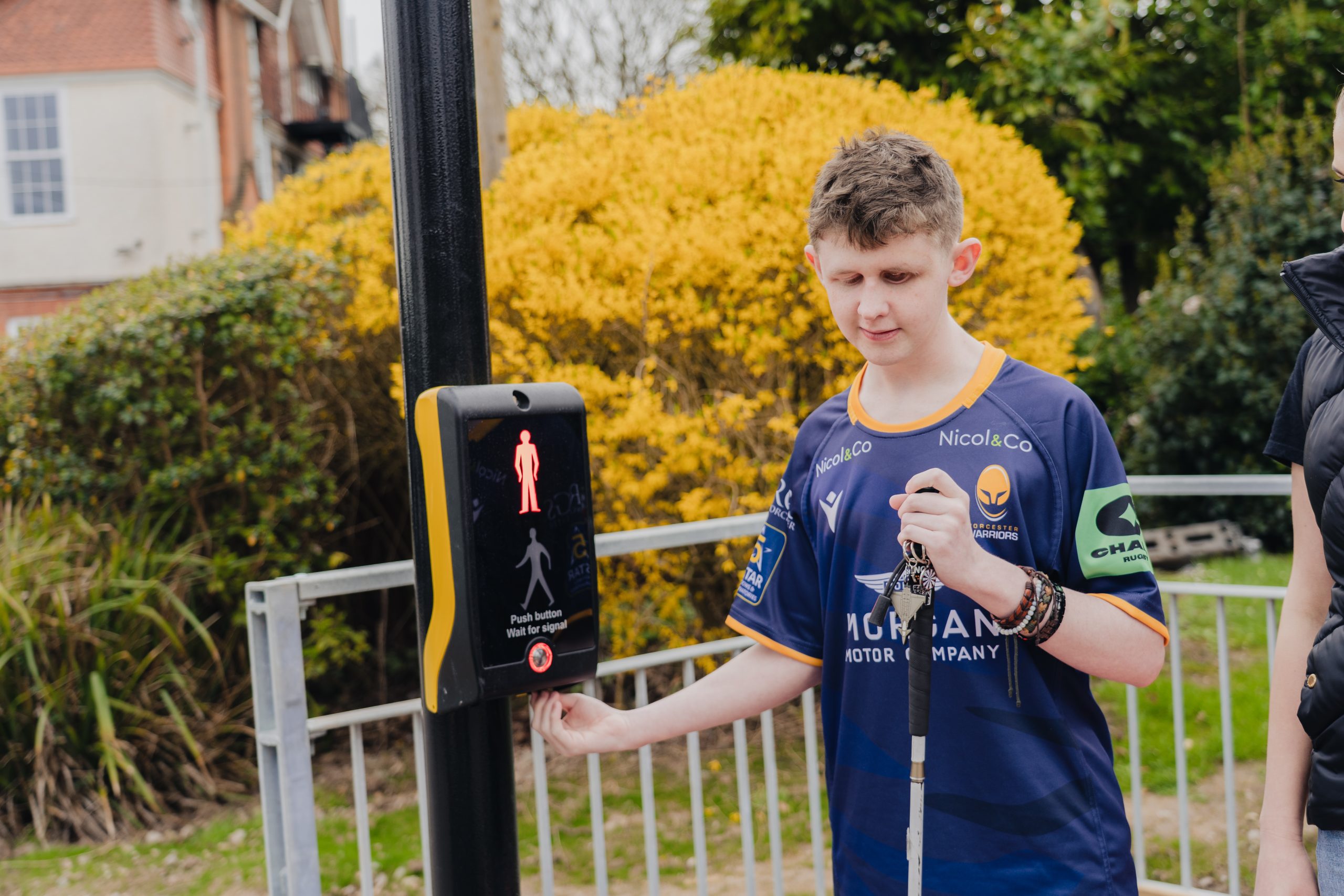 A student standing at a pedestrian crossing presses the button on the signal box with one hand while holding a white cane in the other. The signal box displays a red standing figure indicating not to cross. Behind the person is a metal railing, green shrubbery, and a large yellow bush. A building with red brick and white window frames is visible to the left.