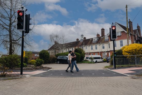Two people walk together across a pedestrian crossing while the traffic lights on both sides display red signals for vehicles. The crossing area is bordered by low shrubs, trees, and a metal railing on the right. In the background, there are several parked cars, a small bus, and a row of older-style buildings with chimneys and sloped roofs under a partly cloudy sky.