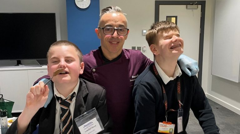 Twostudents are seated together at a table with a chocolatier standing behind them.