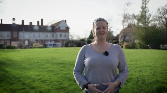 Naomi Riches standing on the front lawn at NCW, wearing a light grey top with a small microphone clipped to it. The school buildings and trees are visible in the background under a bright sky