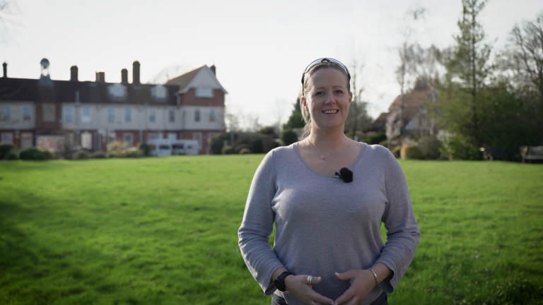 Naomi Riches standing on the front lawn at NCW, wearing a light grey top with a small microphone clipped to it. The school buildings and trees are visible in the background under a bright sky