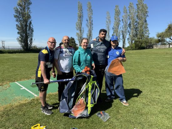 A group of adults from New College Worcester, UK Blind Baseball and international blind baseball partners stand together on a grassy field at NCW, with tall trees and blue sky in the background. They are holding blind baseball equipment including a bat, balls and training accessories.