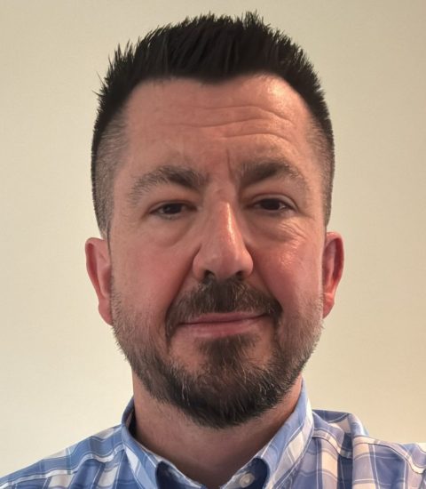 Head shot of Peter Hylton smiling at the camera, he is wearing a blue and white checkered shirt.