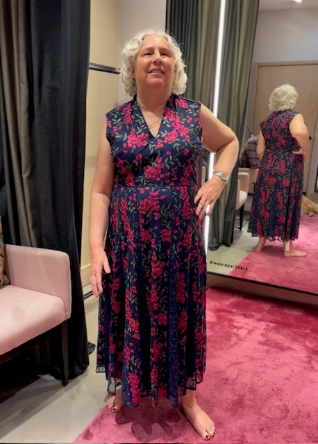 A person stands on a pink carpet in a changing room, trying on a sleeveless navy dress with a pink floral pattern. The person is barefoot and holding a white cane in one hand. Behind them is a full-length mirror showing the back of the dress, with dark curtains and a chair visible in the fitting area.