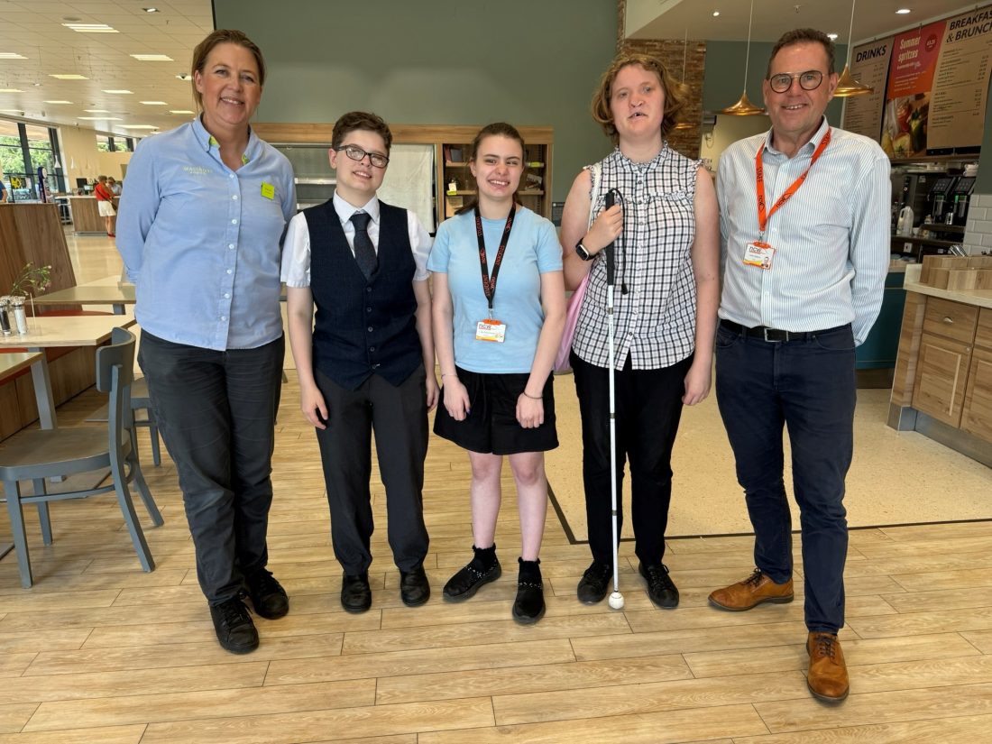 Five people stand together in the café in Waitrose Worcester during a work experience placement. Two adults stand at either end of the group, with three young people in the centre. One student is holding a long cane. Several members of the group are wearing lanyards. Tables, chairs, and a service counter with menu boards are visible in the background. The group is facing the camera in a well-lit indoor setting.