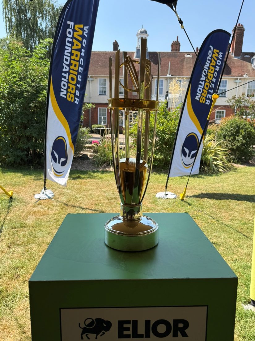 The Elior Championship Rugby Trophy stands on display outdoors on a black plinth. Worcester Warriors Foundation flags are positioned on either side of the trophy, with school buildings and landscaped gardens visible in the background on a sunny summer day.