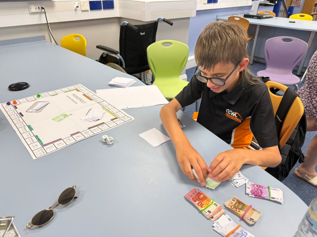 Student explores a Braille version of Monopoly in French, reading Braille cards and game materials while seated at a table.