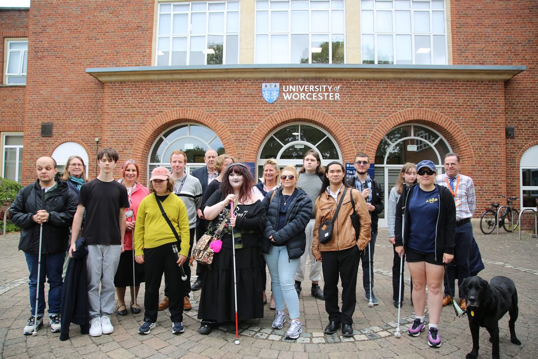 Group photo of students and staff from New College Worcester (NCW) and the Royal National College for the Blind (RNC) standing outside a University of Worcester campus building. Several people are holding long canes, and a guide dog is standing at the front of the group. The University of Worcester logo is visible above the entrance behind them as they take part in a Visual Impairment Information Day focused on preparing for higher education.