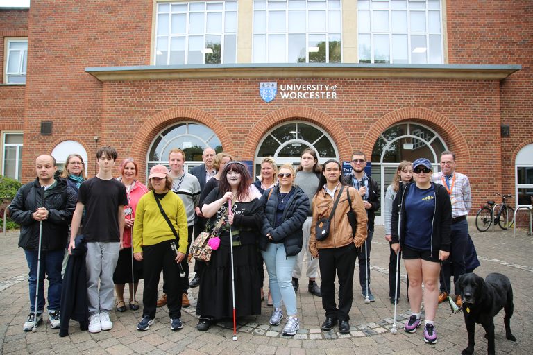 Group photo of students and staff from New College Worcester (NCW) and the Royal National College for the Blind (RNC) standing outside a University of Worcester campus building. Several people are holding long canes, and a guide dog is standing at the front of the group. The University of Worcester logo is visible above the entrance behind them as they take part in a Visual Impairment Information Day focused on preparing for higher education.