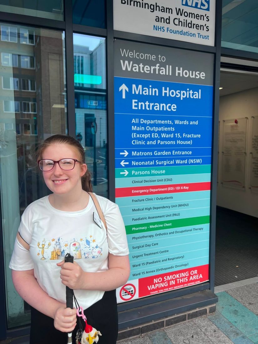 Photo of Emma smiling outside of the hospital, she is holding her cane