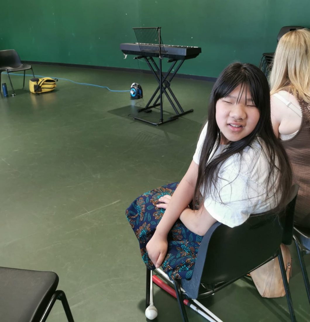 Keziah smiling at the camera, she is in a large room with a keyboard and a blonde person is sat next to her