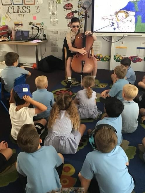 Len playing the cello to the class, the children are facing him and only theirs heads visible