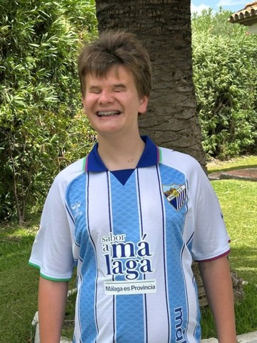 A person stands outdoors in a grassy area beside a tree, wearing a blue and white striped football shirt with the Málaga CF crest on the front. Shrubs and garden plants can be seen in the background, and the photo was taken on a bright, sunny day