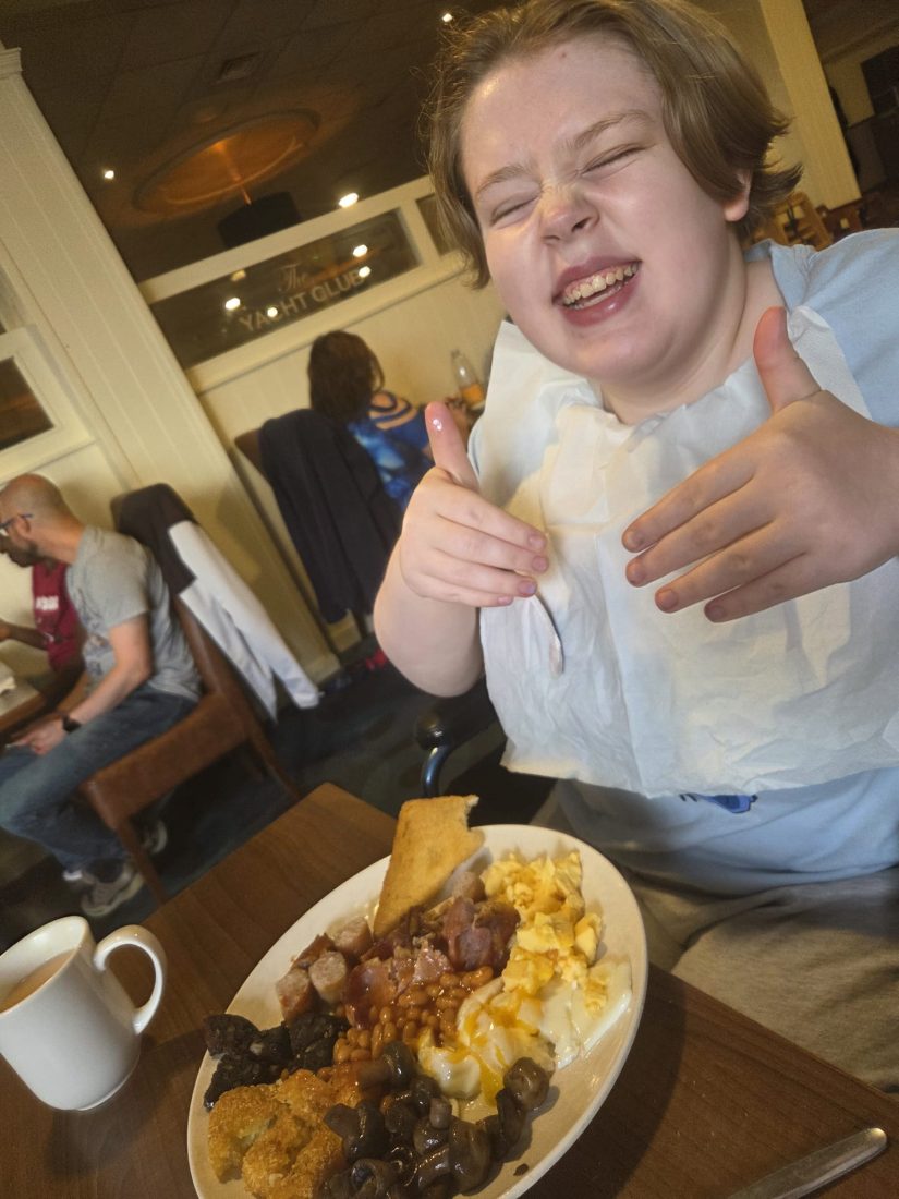 Kyla sits at a restaurant table with a large cooked breakfast in front of them that includes eggs, bacon, sausages, beans, mushrooms, black pudding and toast. A mug of tea sits on the table beside the plate. The person is making hand gestures towards the camera. Other diners, tables and chairs can be seen in the restaurant background, and a sign reading “The Yacht Club” is visible on the wall. Provide your feedback on BizChat Sources
