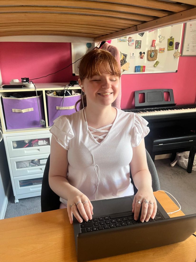 Natalie sits at a desk using a laptop, with earphones connected and her hands resting on the keyboard. Behind her is a digital piano, storage units with purple fabric drawers, and a noticeboard displaying photographs, certificates and mementos. The room has pink walls and the underside of a bunk bed is visible above the desk.