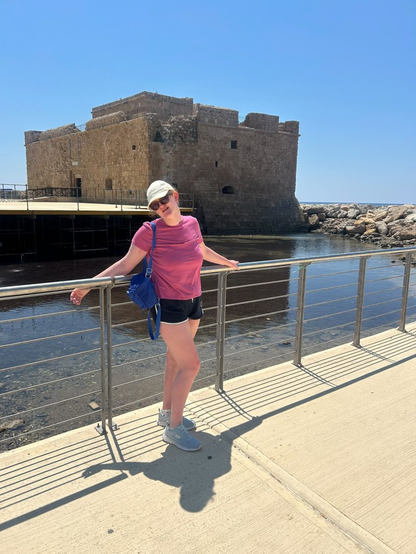 Liv stands on a waterfront walkway, leaning against a metal railing beside a calm body of water. Liv is wearing a pink T-shirt, dark shorts, trainers, sunglasses and a light-coloured cap, with a blue shoulder bag. Behind Liv is a large historic stone fort beside the water, with rocks and a clear blue sky in the background on a bright, sunny day.