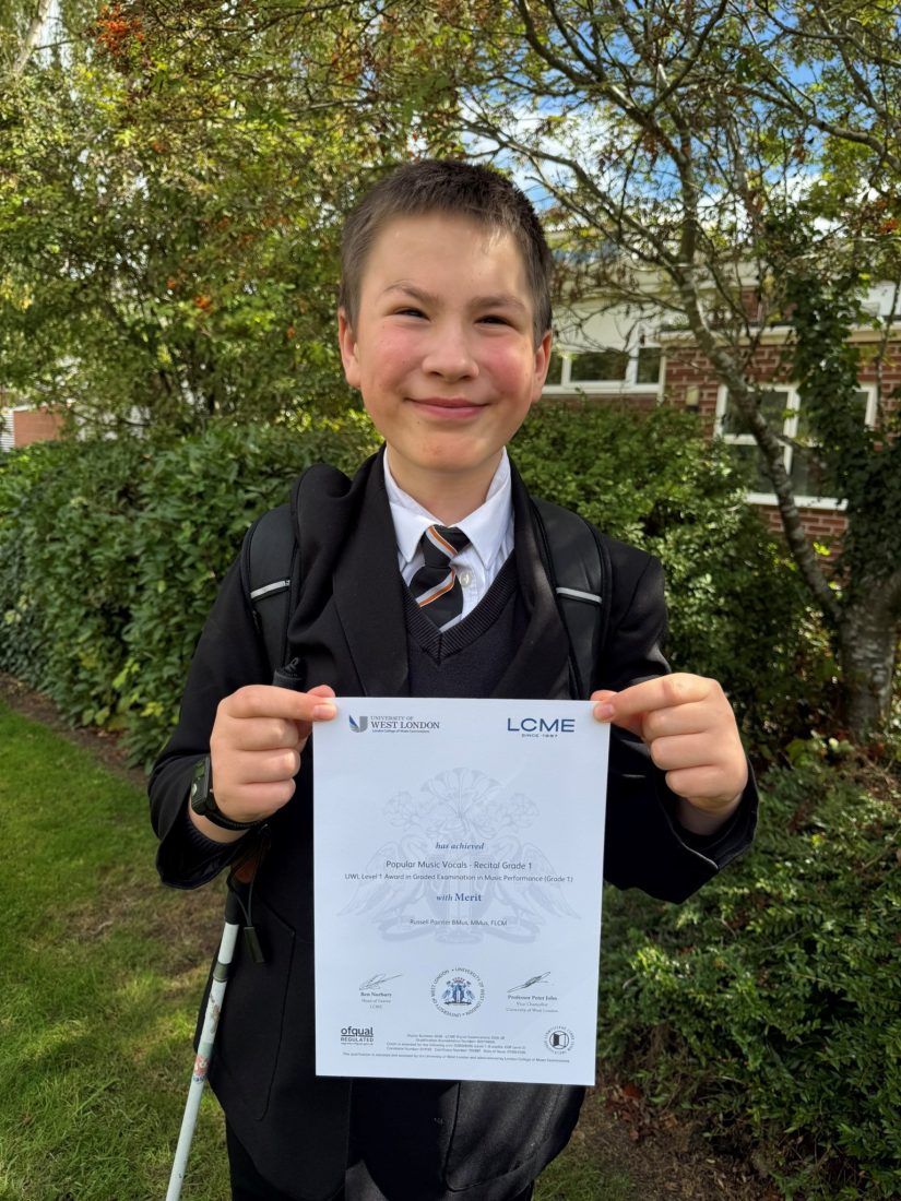 Ed stands outdoors in front of trees and greenery, holding an LCME (London College of Music Examinations) certificate with both hands. Ed is wearing a school blazer, jumper, shirt and striped tie, with a backpack over one shoulder and a long white cane visible at his side. The certificate shows a Merit in Popular Music Vocals Recital Grade 1. A brick school building can be seen in the background, and the photograph is taken in bright sunshine on the NCW campus.