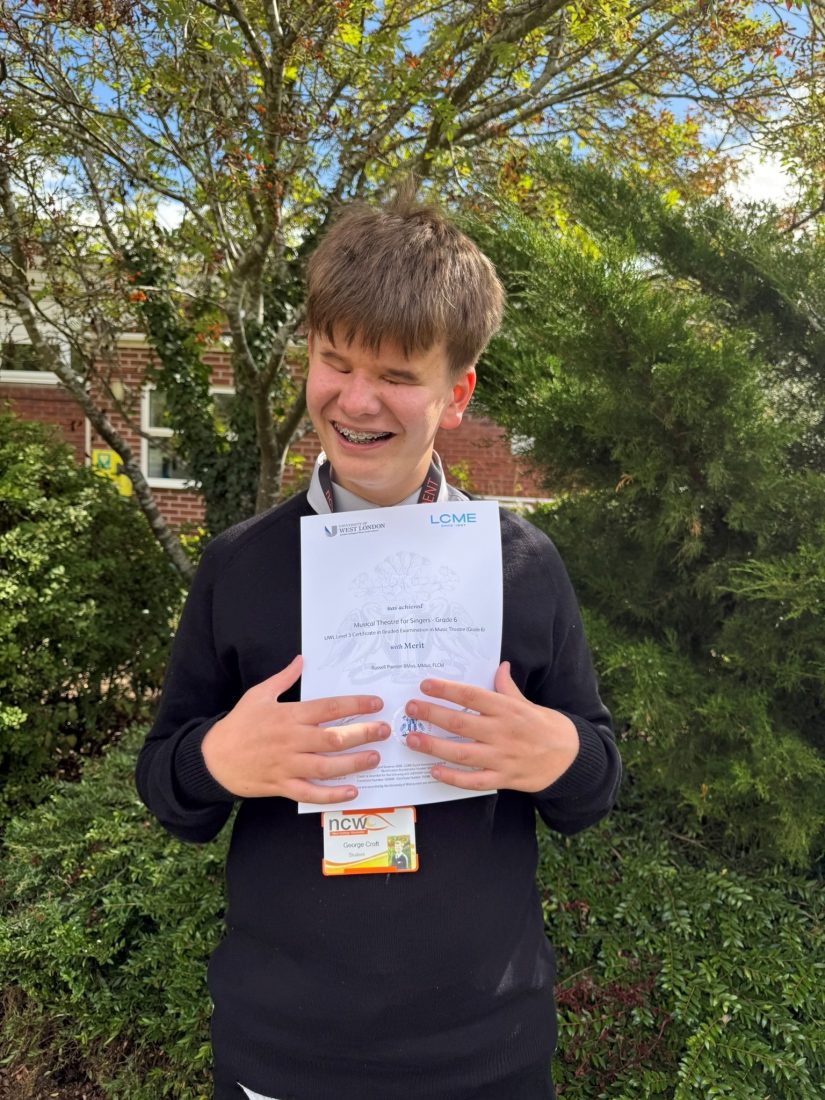 George stands outdoors in front of trees, shrubs and a brick building, holding an LCME (London College of Music Examinations) certificate against his chest. He is wearing a dark jumper and an NCW student lanyard. The certificate shows a Merit in Musical Theatre for Singers Grade 6. The photograph was taken on the NCW campus in bright sunshine, with greenery and autumn berries visible in the background.