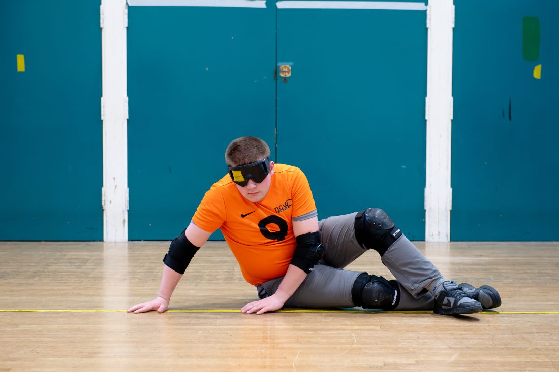 A student takes part in a goalball activity inside a sports hall. The student is seated on a wooden sports floor and wears a bright orange NCW sports shirt, protective elbow pads, knee pads and blackout goggles used in goalball. One hand is placed on the floor while the other supports their movement as they position themselves near a court line. Large blue wall panels form the background, and the image highlights the specialist equipment and techniques used in this vision impairment-inclusive sport.