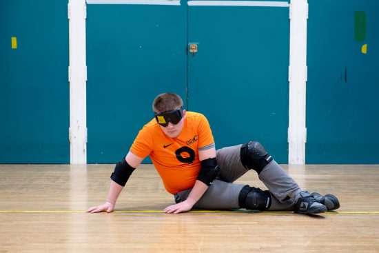 A student takes part in a goalball activity inside a sports hall. The student is seated on a wooden sports floor and wears a bright orange NCW sports shirt, protective elbow pads, knee pads and blackout goggles used in goalball. One hand is placed on the floor while the other supports their movement as they position themselves near a court line. Large blue wall panels form the background, and the image highlights the specialist equipment and techniques used in this vision impairment-inclusive sport.