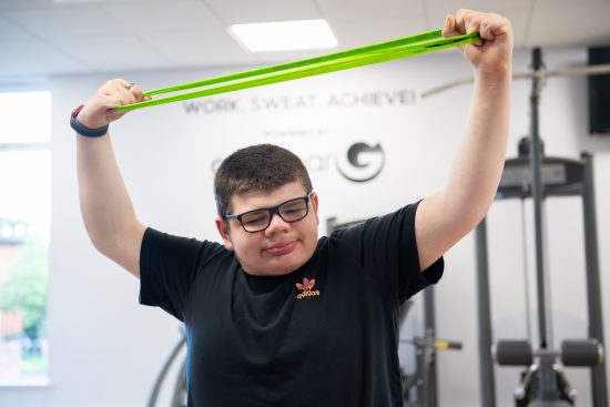 A student takes part in an exercise session in a fitness suite, stretching a bright green resistance band above their head with both hands. The student is wearing glasses, a black Adidas t-shirt and a smartwatch. Gym equipment can be seen in the background, and the words "WORK SWEAT ACHIEVE" are displayed on the wall behind them. The photograph is taken indoors in a well-lit exercise space at NCW.