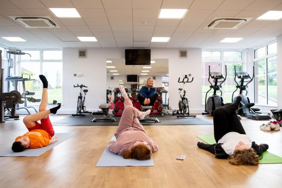 Three students take part in a fitness session in NCW's gym. The students are lying on exercise mats on a wooden floor, each raising one leg as part of a guided workout. An instructor sits on a bench at the front of the room, leading the session. The bright, modern fitness suite contains exercise bikes, weights, benches and cardio equipment, with large windows allowing natural light into the space. A wall mirror reflects the gym equipment and instructor, highlighting the spacious workout environment.