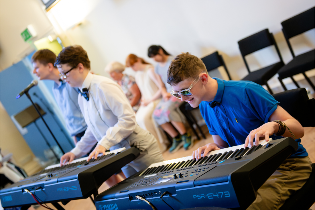 Two students perform on electronic keyboards during a music event. One student in a blue polo shirt plays a Yamaha keyboard in the foreground, while another student in a white shirt and bow tie plays a second keyboard beside a microphone. Audience members are seated in the background watching the performance.