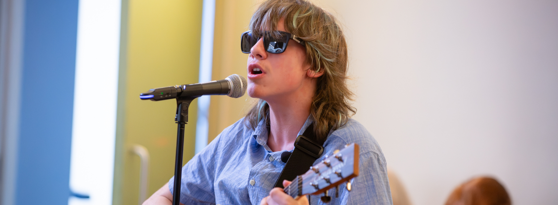A student performs with an acoustic guitar while singing into a microphone. The student is wearing dark glasses and a light blue shirt and is standing in a bright indoor space. Several audience members can be seen seated in the background, slightly out of focus, watching the performance. The image captures the student mid-song during a live music performance.