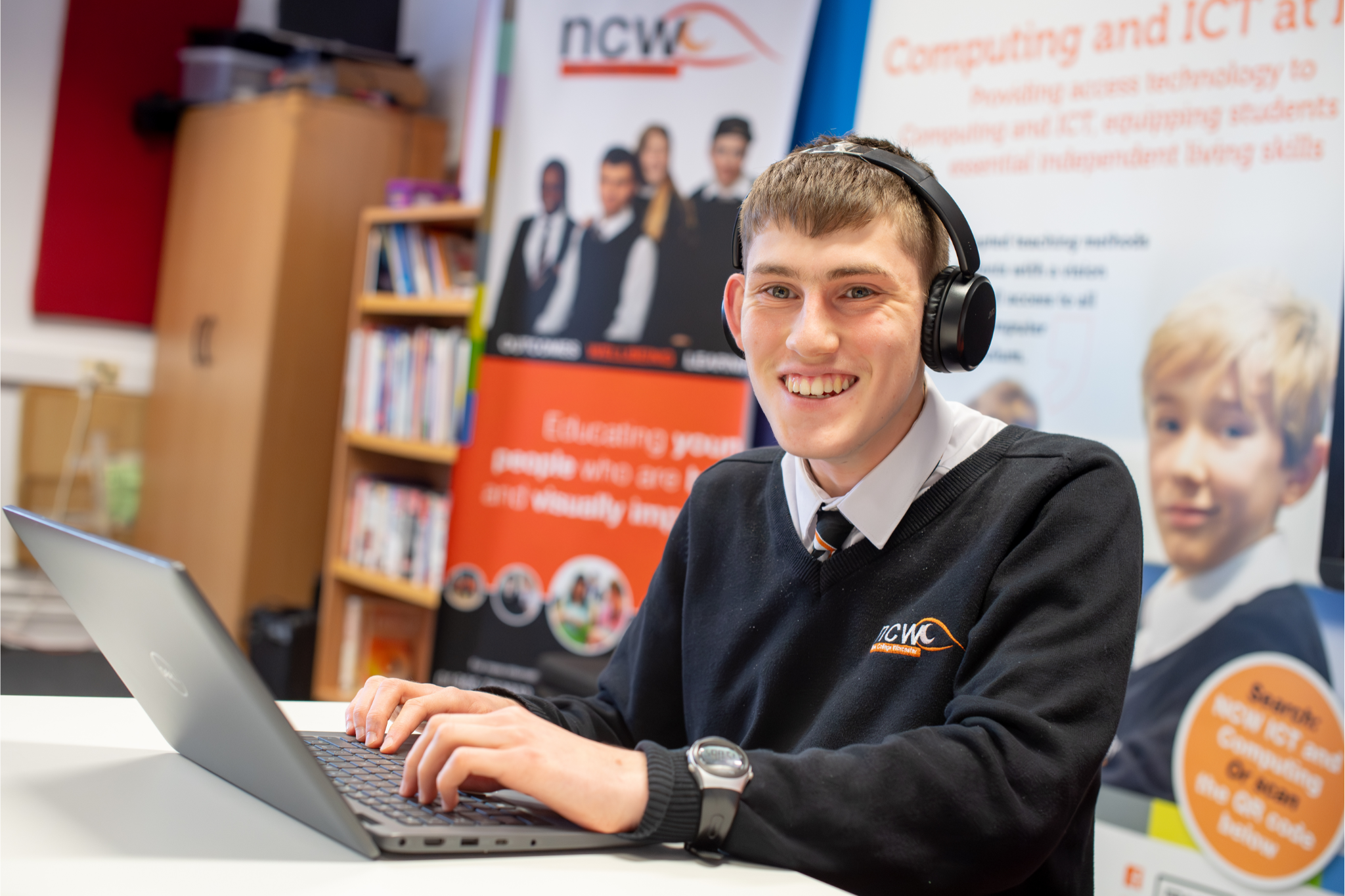 A smiling student in uniform wearing headphone and working on a laptop in a classroom