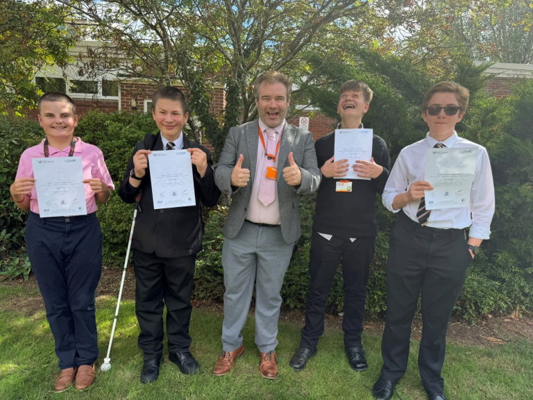 Students and Mr Painter stand outdoors on a lawn in front of trees and a brick building, each holding a certificate. Four students stand alongside a member of staff in a grey suit and pink tie, who is giving two thumbs up. One student is holding a long white cane. The group is arranged in a line and posing for a celebratory photograph in bright sunshine following the presentation of singing exam results. The certificates are from LCME (London College of Music Examinations).